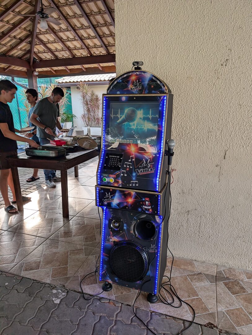 Máquina de karaokê DanSan montada em uma área de churrasco, com convidados preparando a comida ao fundo